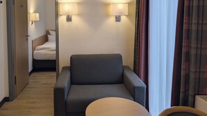 A hotel room with a grey armchair, a round wooden table, and a bed with white linens. There are two wall-mounted lamps on either side of the bed. - Garner Hotel Hamburg Wandsbek Marktplatz by IHG (Hamburg)