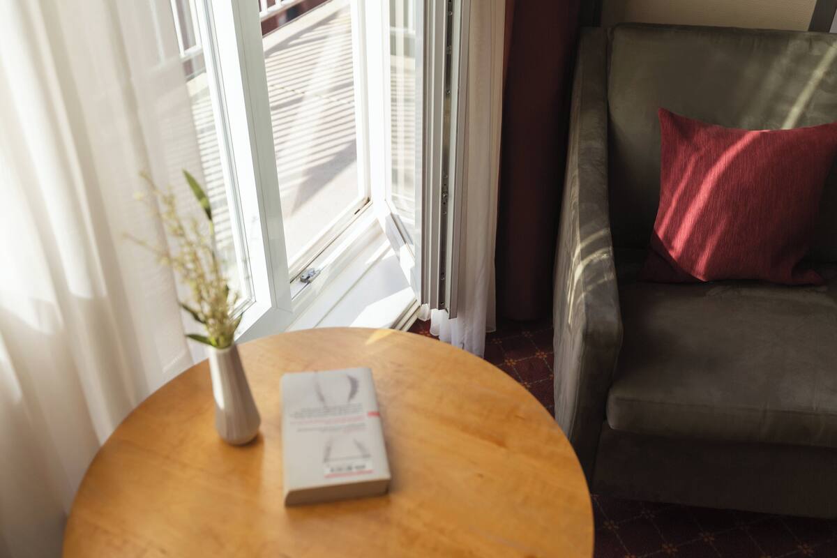 A round wooden table with a white vase and a book, a grey sofa with a red pillow, and a window with blinds.
