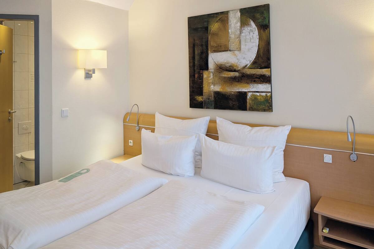 A hotel room with a double bed, a nightstand, a wall-mounted lamp, and a painting.
