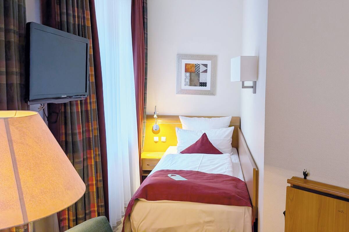 A hotel room with a bed, a television mounted on the wall, a bedside table with a lamp, and a wall-mounted light fixture.