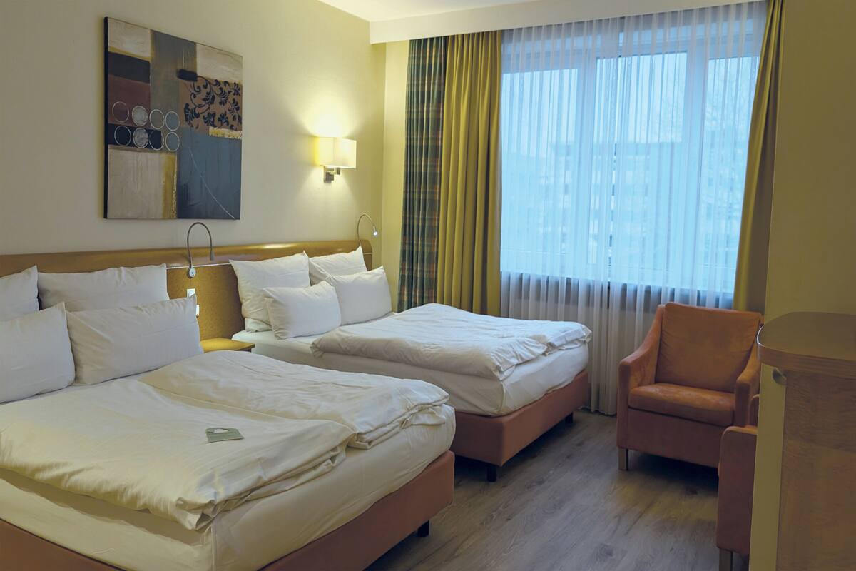 A hotel room with two beds, a chair, a lamp, and a painting on the wall.