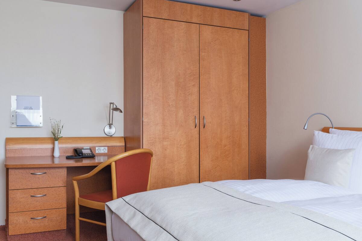 A hotel room with a bed, a desk with drawers, a chair, a wardrobe, and a bedside table with a lamp.