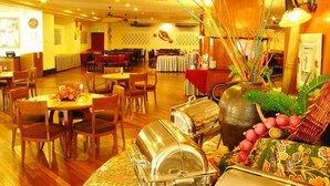 Restaurante com mesas e cadeiras de madeira, estações de buffet e um vaso decorativo com plantas.