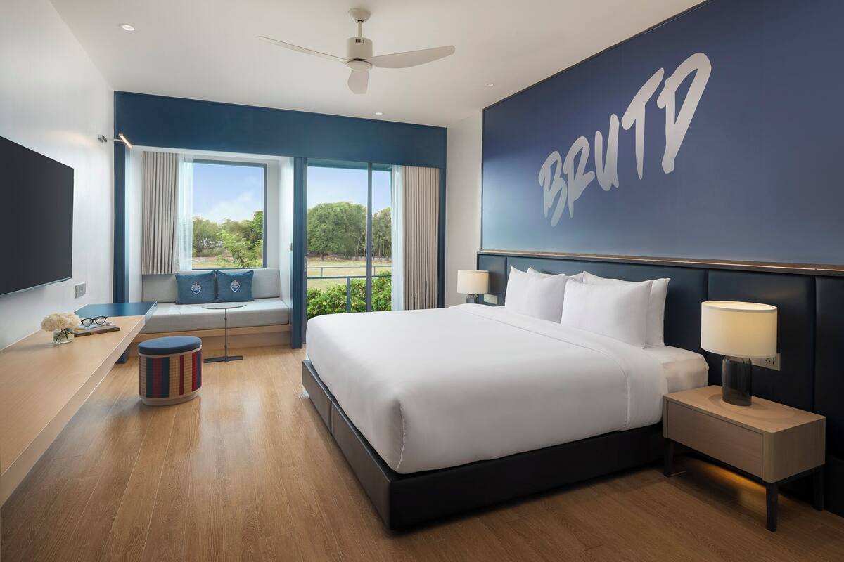 Habitación de hotel moderna con una cama grande, un escritorio, una silla, una lámpara de pie y vistas al exterior.