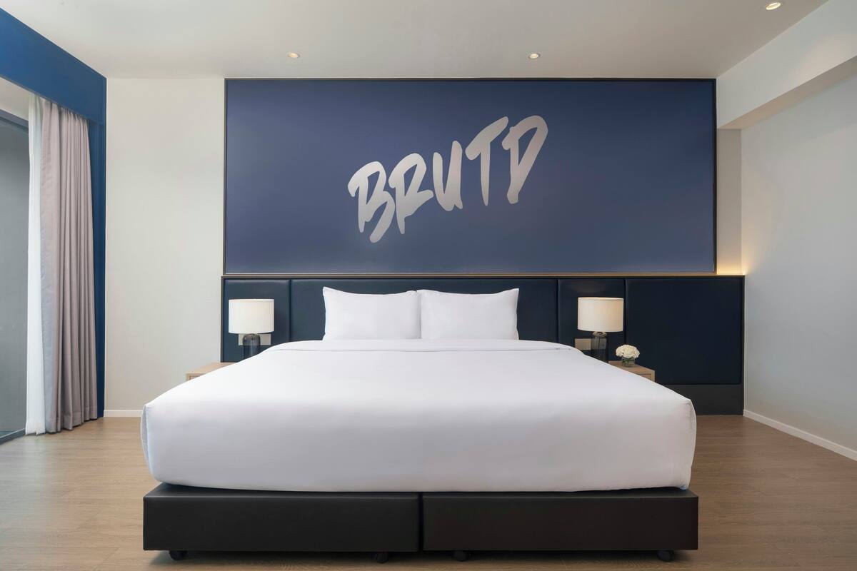 A modern hotel room with a large bed, two bedside tables with lamps, and a wall-mounted screen displaying the word 'BRUTD'.