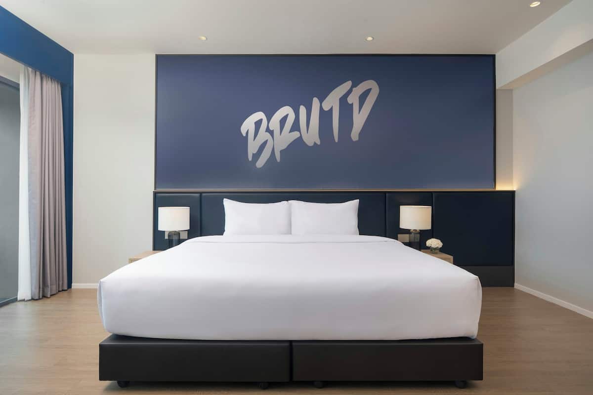 Ein modernes Hotelzimmer mit einem groĂen Bett, zwei Nachttischen mit Lampen und einem an der Wand montierten Bildschirm, der das Wort 'BRUTD' anzeigt.