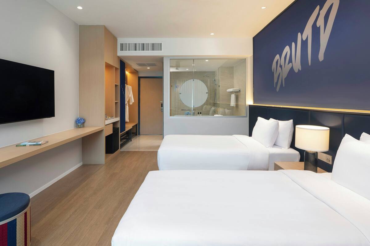 A hotel room with two beds, a TV, a desk, a mirror, and a large wall with the name 'BRUTIO'.