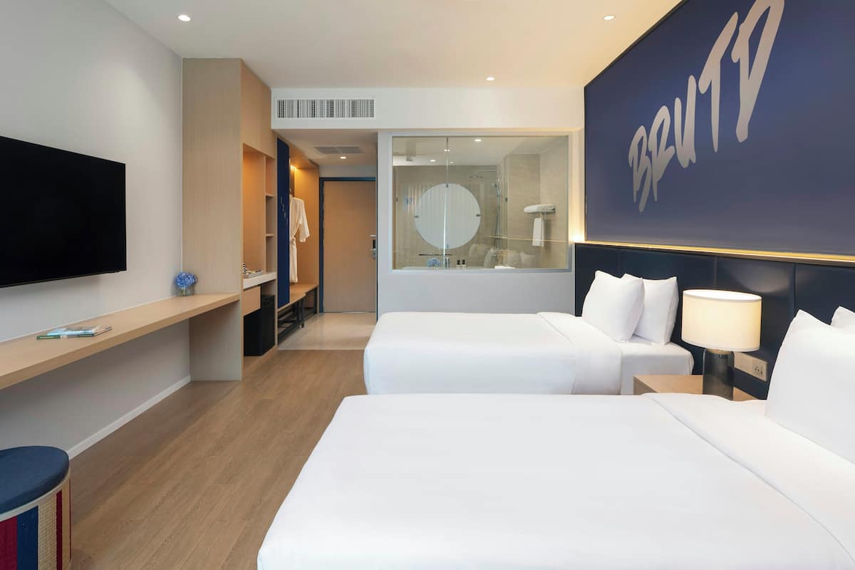 A hotel room with two beds, a TV, a desk, a mirror, and a large wall with the name 'BRUTIO'.