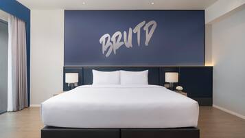 A modern hotel room with a large bed, two bedside lamps, a dark headboard, and a large wall-mounted screen displaying 'BRUTD'.