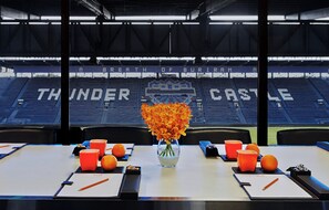 Una mesa de conferencias con un jarrón de flores, tazas anaranjadas y vista a un estadio con la inscripción 'Thunder Castle' en los asientos.