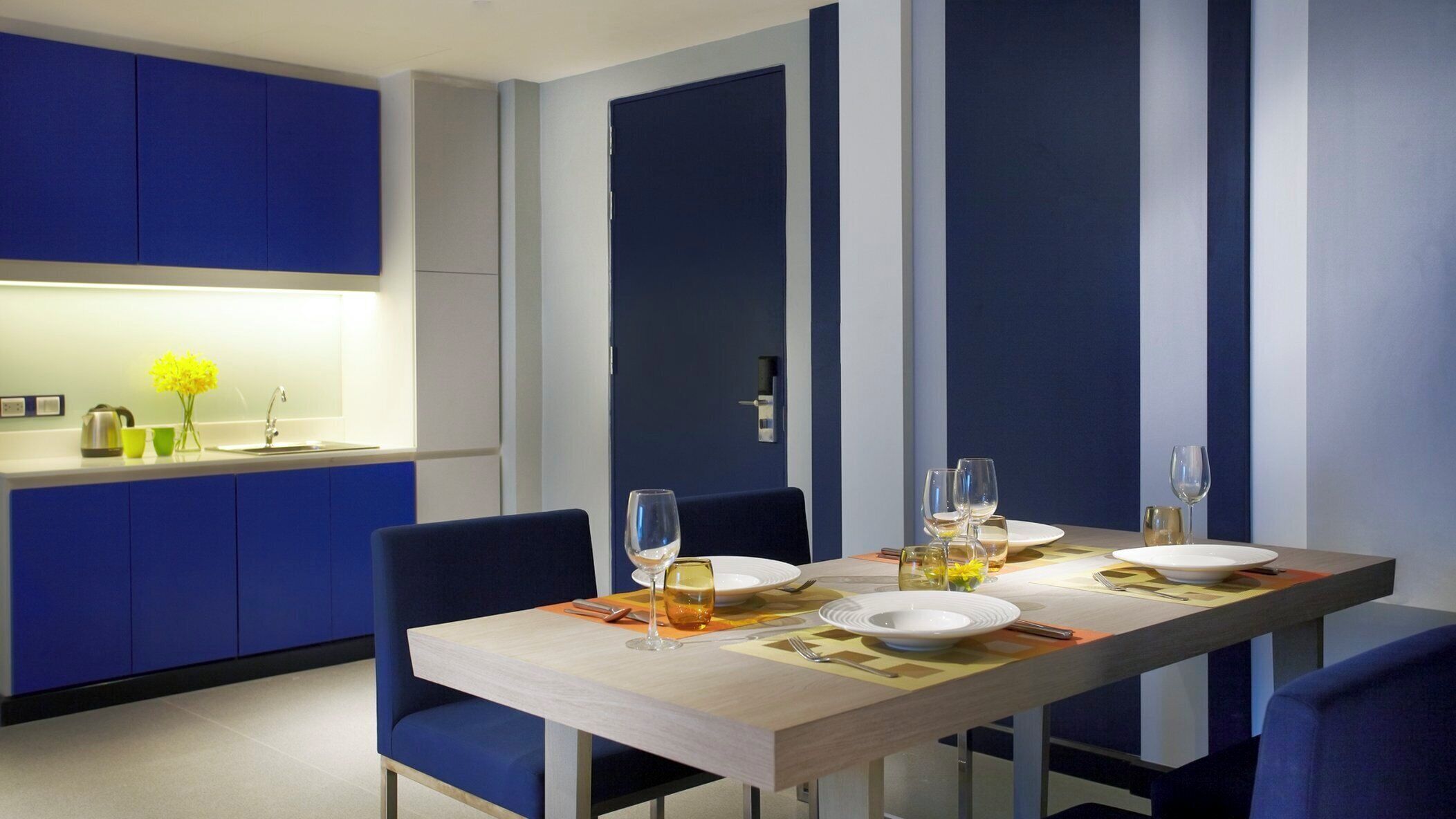 A modern dining area with a rectangular table set for four, blue upholstered chairs, and a kitchen area with blue cabinets and a white countertop.