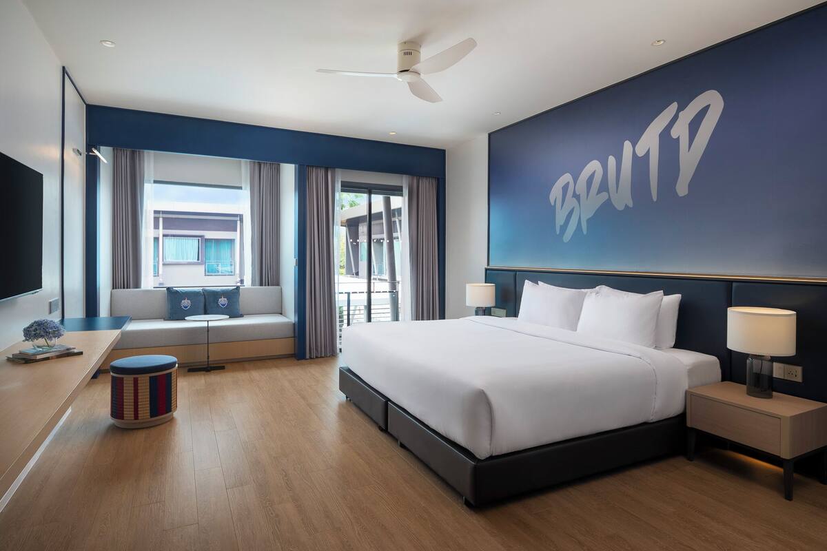 A hotel room with a bed, bedside tables, a desk, a chair, a window with curtains, a ceiling fan, and a wall with the hotel's name.