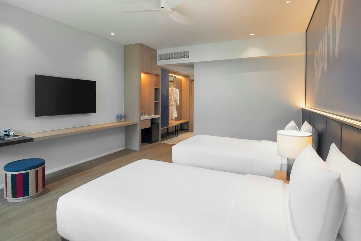 A hotel room with two beds, a flat-screen TV, a ceiling fan, a bedside table with a lamp, and a walk-in closet.