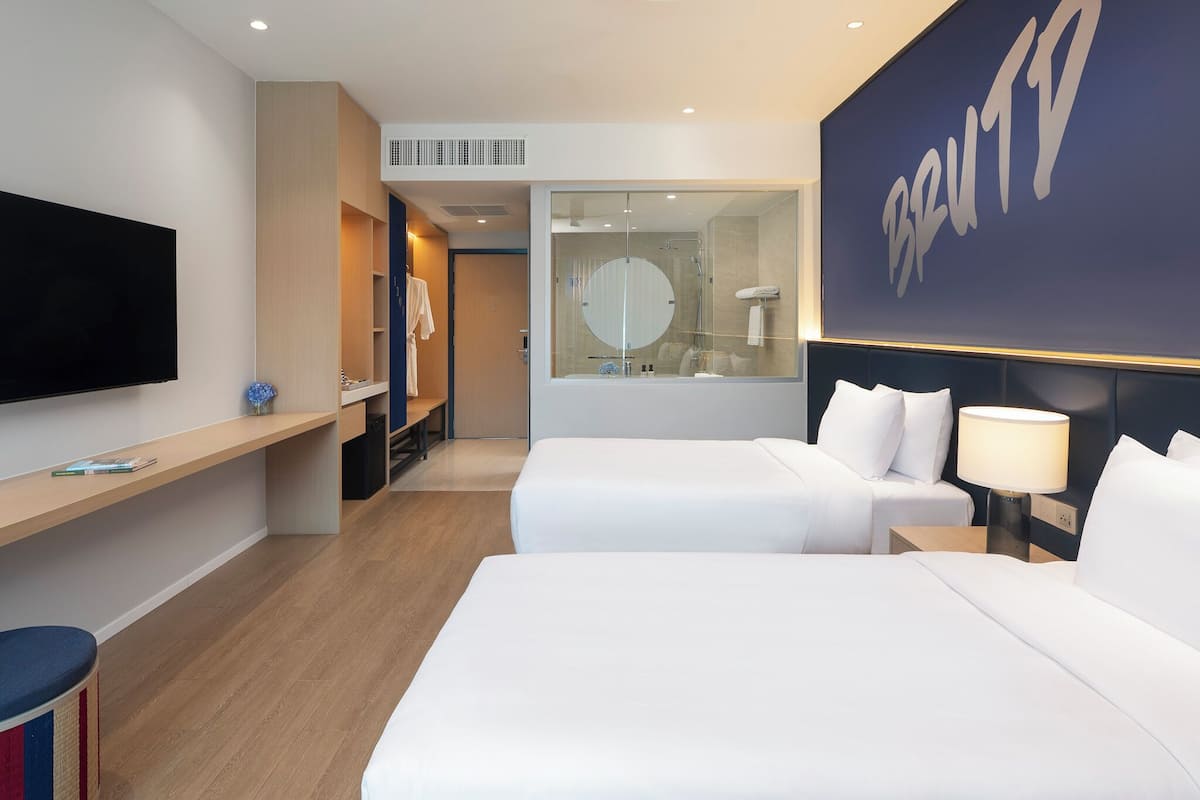 A hotel room with two beds, a TV, a desk, and a bathroom with a large mirror.