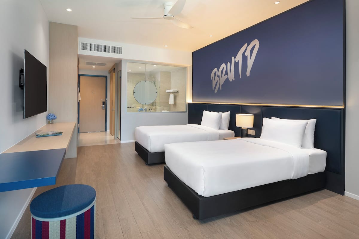A hotel room with two beds, a TV, a desk, and a large wall with the word 'BRUTD' on it.