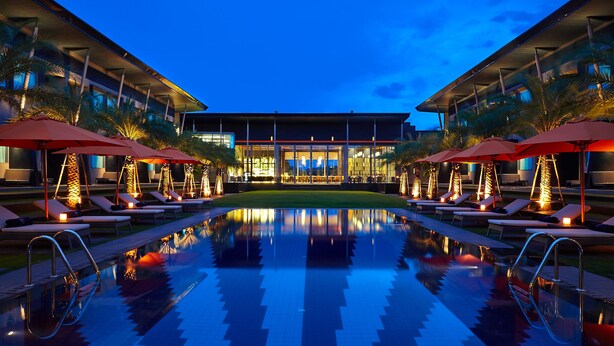 Un área de piscina con sillones de descanso y sombrillas, un estanque reflectante y un edificio con grandes ventanales al fondo.