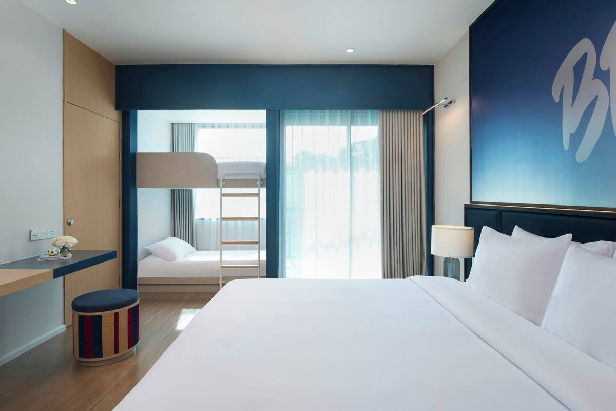 Habitación de hotel con una cama grande, un escritorio, una silla, un taburete pequeño, una ventana con cortinas y una pared con una letra grande.