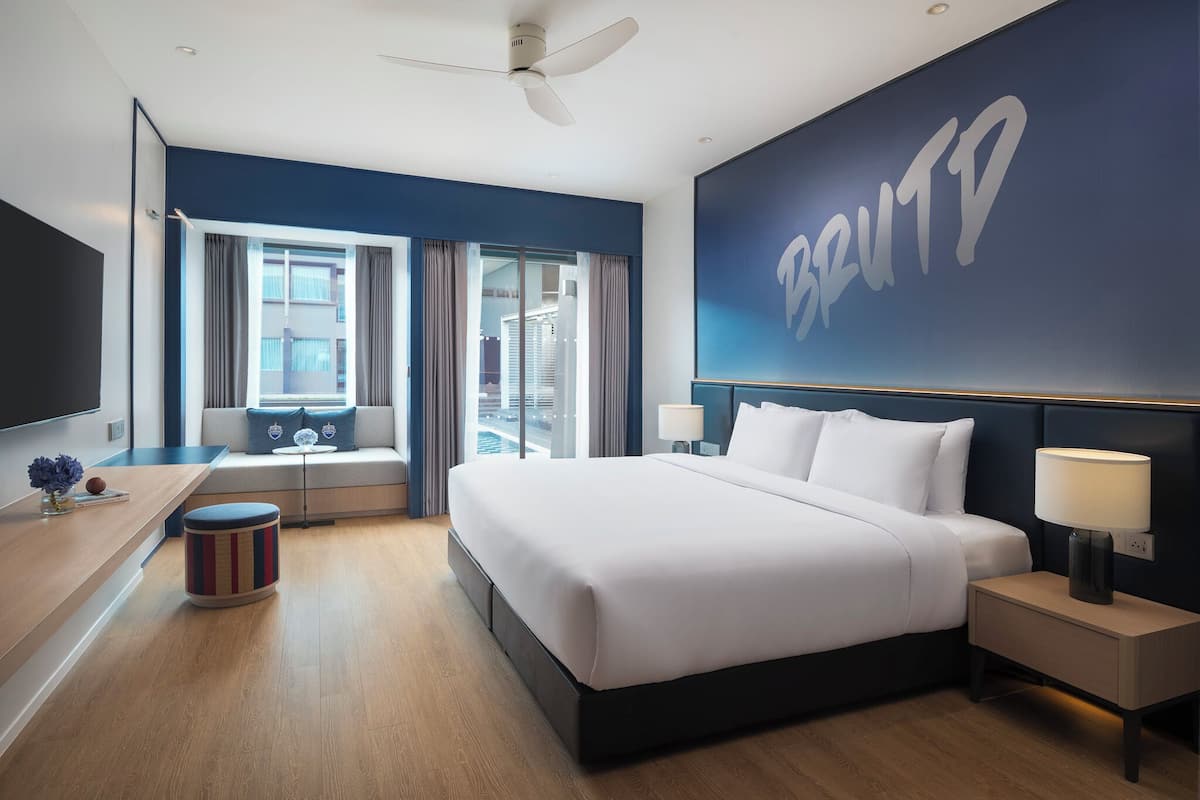 A modern hotel room with a large bed, a flat-screen TV, a sofa, a window with curtains, and a wall with a large logo.
