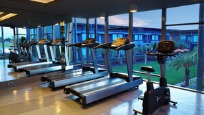 Un gimnasio con cintas de correr y una bicicleta estática, amplias ventanas y vistas a un edificio y zonas verdes al exterior.
