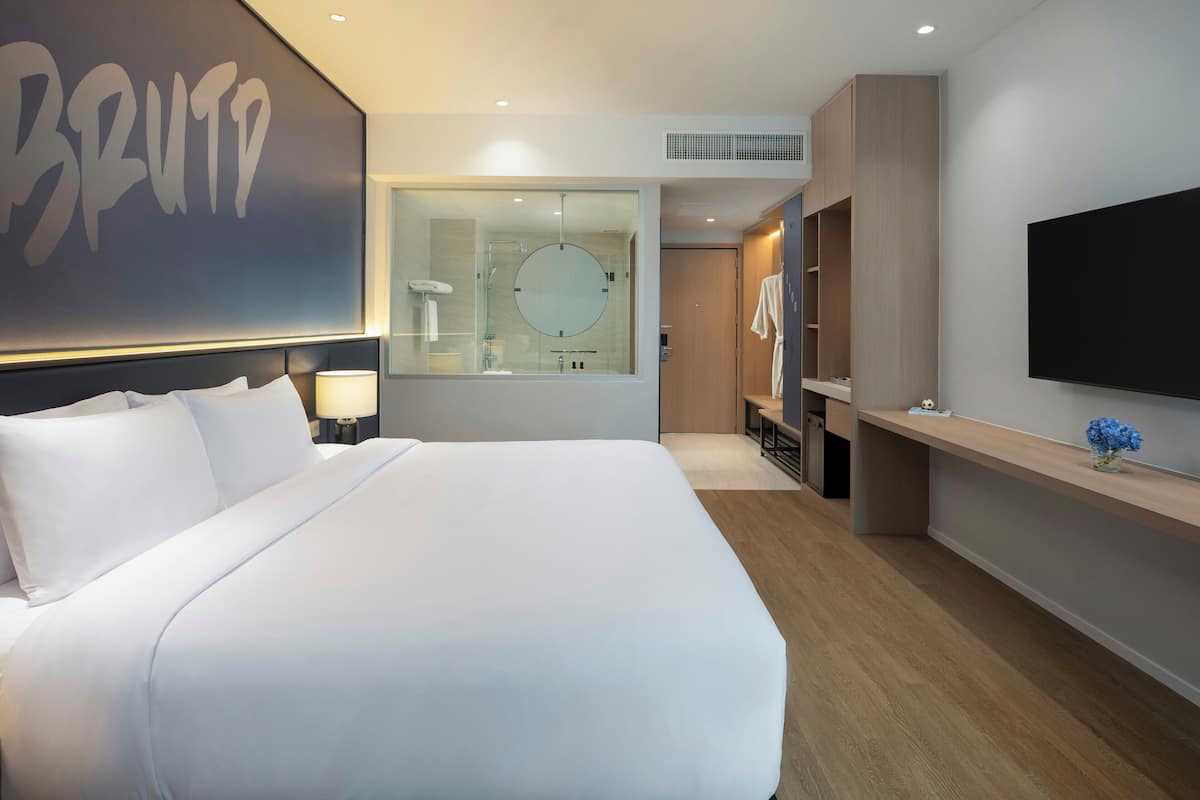 A modern hotel room with a large bed, a TV, a mirror, and a bathroom visible through a glass partition.