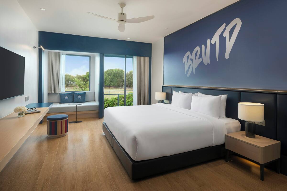 A modern hotel room with a large bed, a desk, a chair, a window with curtains, a ceiling fan, and a wall with a large logo.