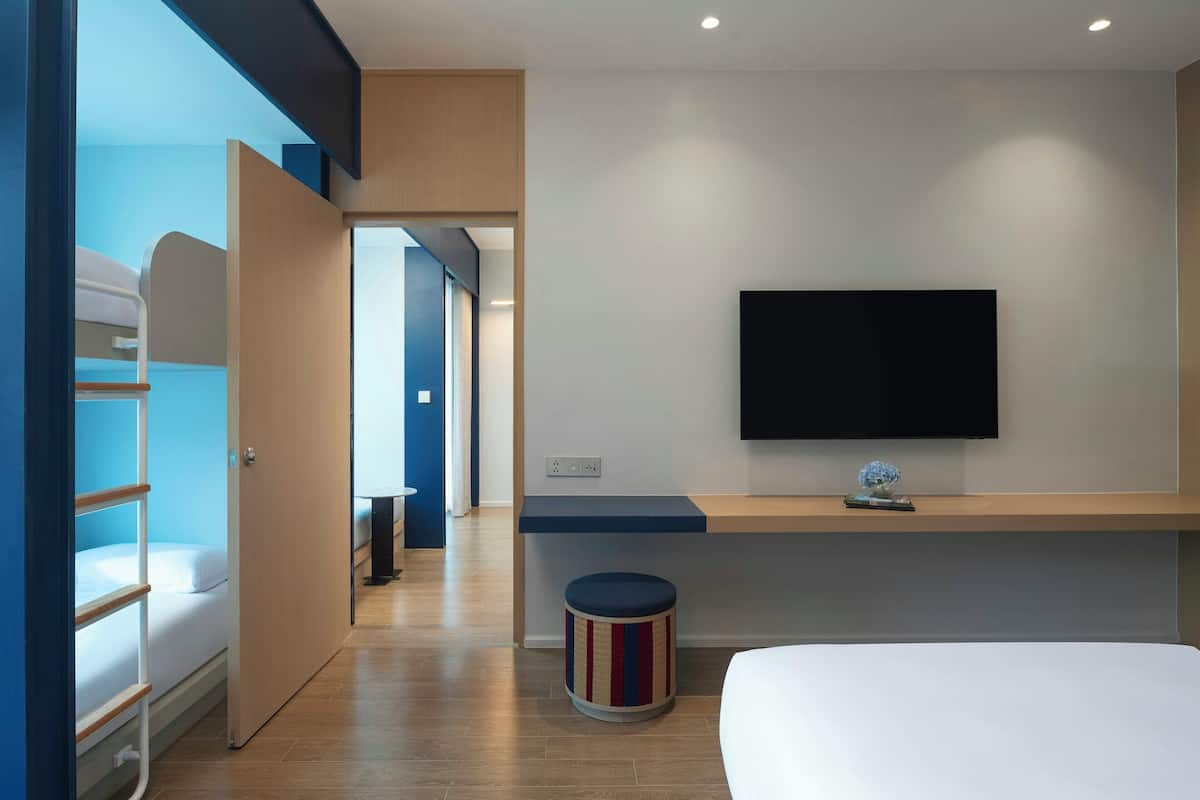 A modern hotel room with a bed, a TV mounted on the wall, a small stool, and a door leading to another room.