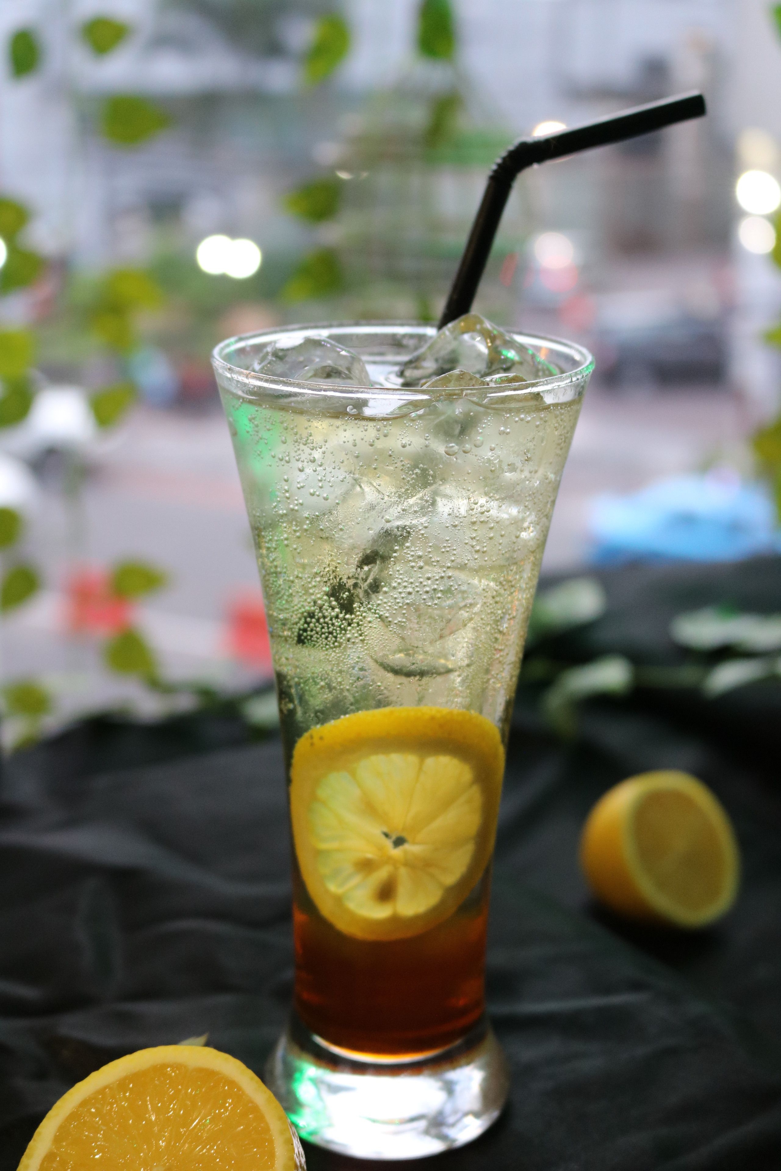 A tall glass with a straw, filled with a beverage and ice cubes, garnished with a slice of lemon, on a dark cloth.