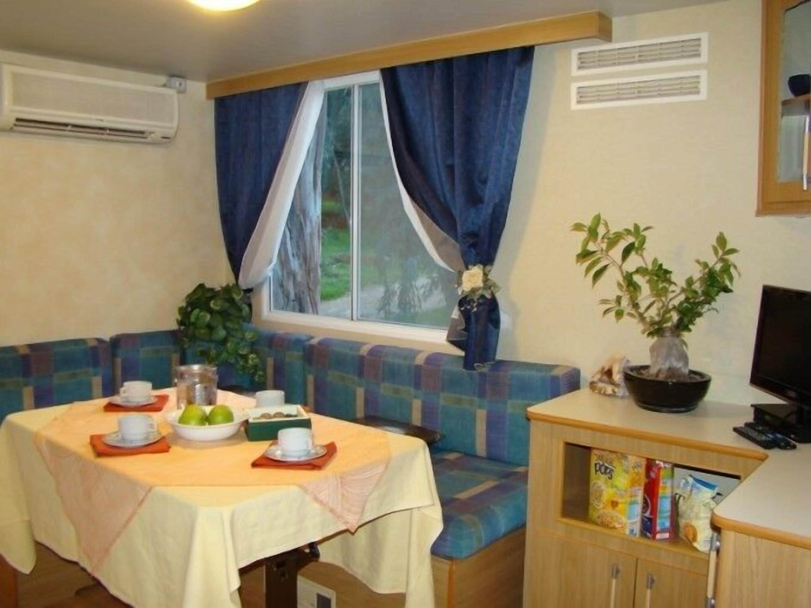 A cozy room with a dining table set for two, a bench, a small cabinet with snacks, a potted plant, and a window with curtains.