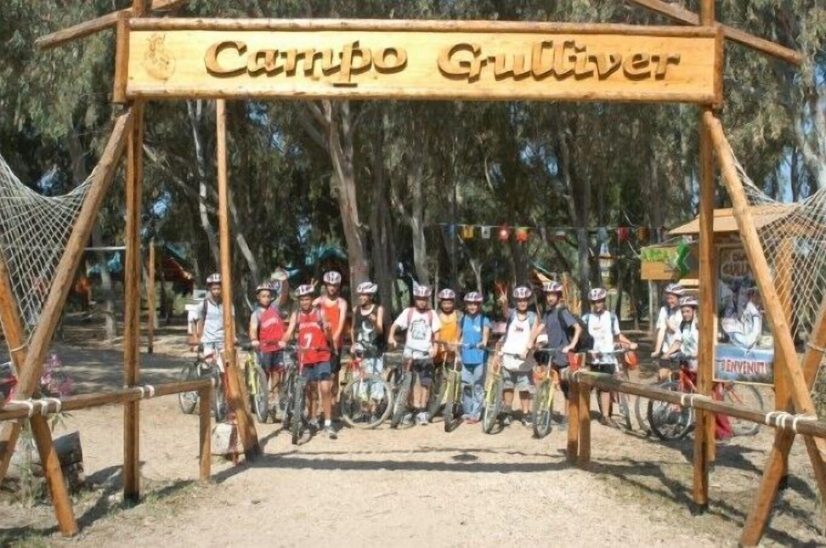 Un letrero de madera con el texto "Campo Grulliver" sobre un grupo de ciclistas.