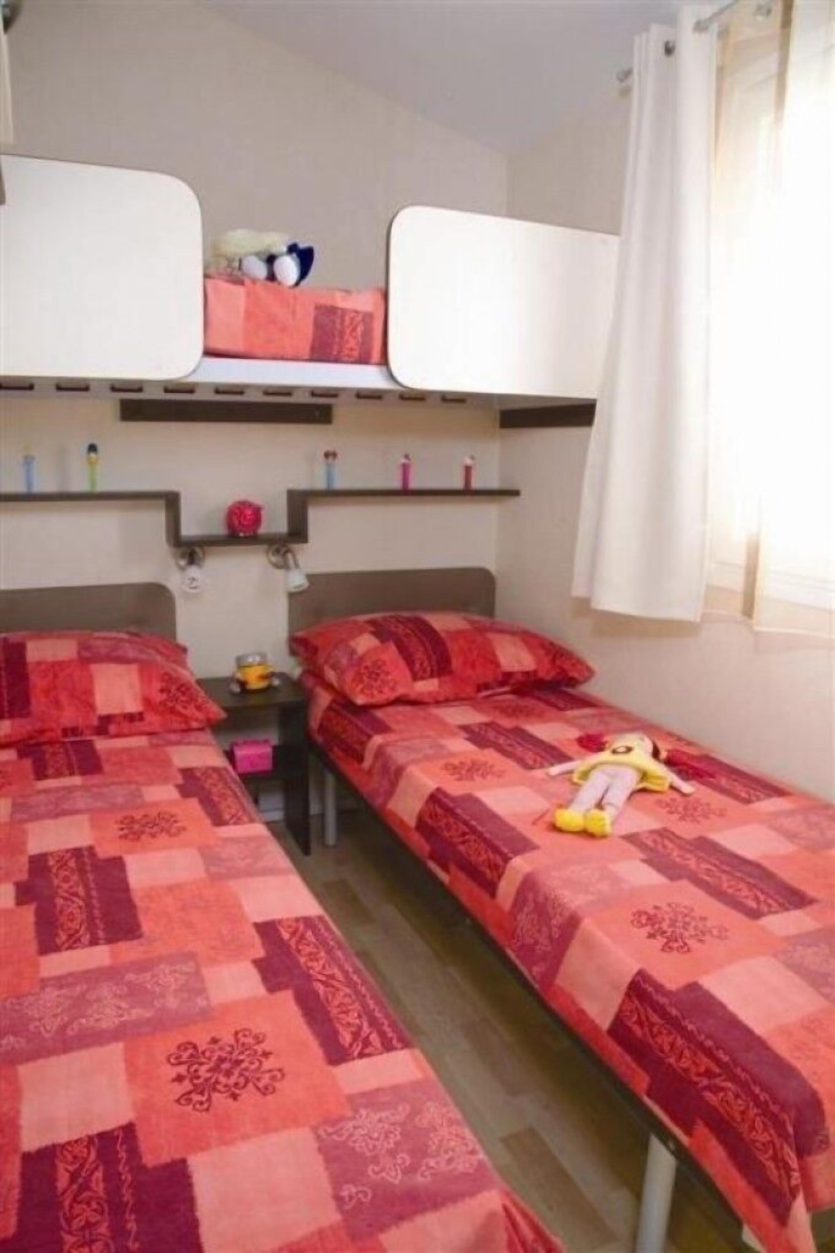 A room with two beds, each with a red patterned bedspread, a nightstand with a lamp, and a shelf above the beds with various items.