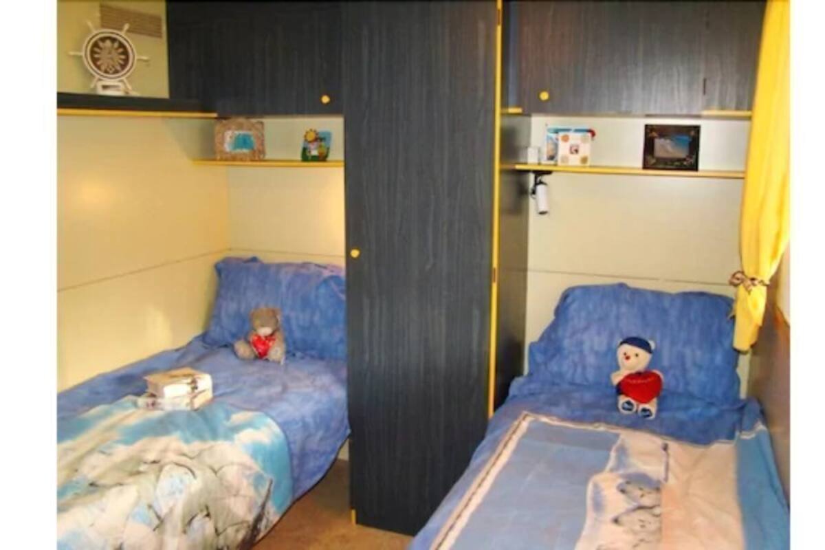A room with two beds, each with a blue bedspread and a teddy bear. There is a shelf with various items and a door with a curtain.