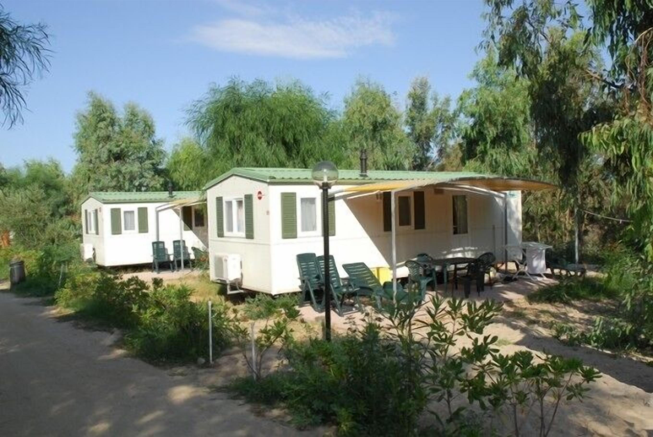 A row of two single-story mobile homes with attached porches, green outdoor seating, and a dirt path leading to the entrance.