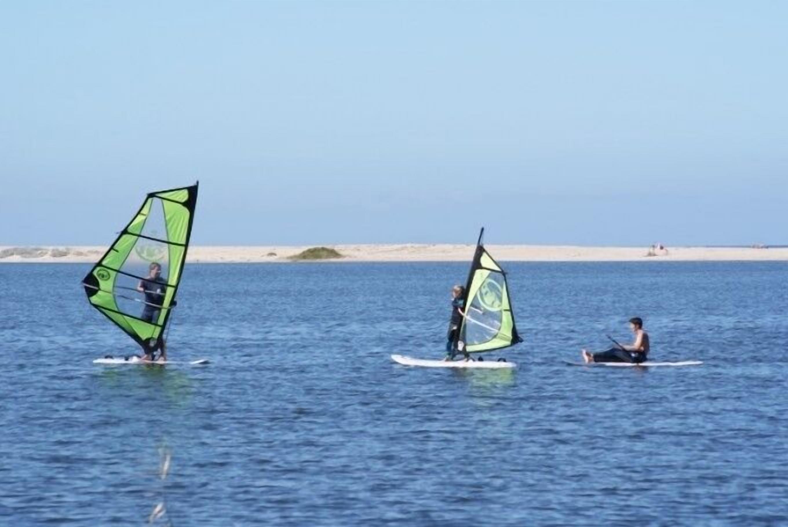 Tres personas practicando windsurf sobre una superficie acuática tranquila, con cielos despejados.