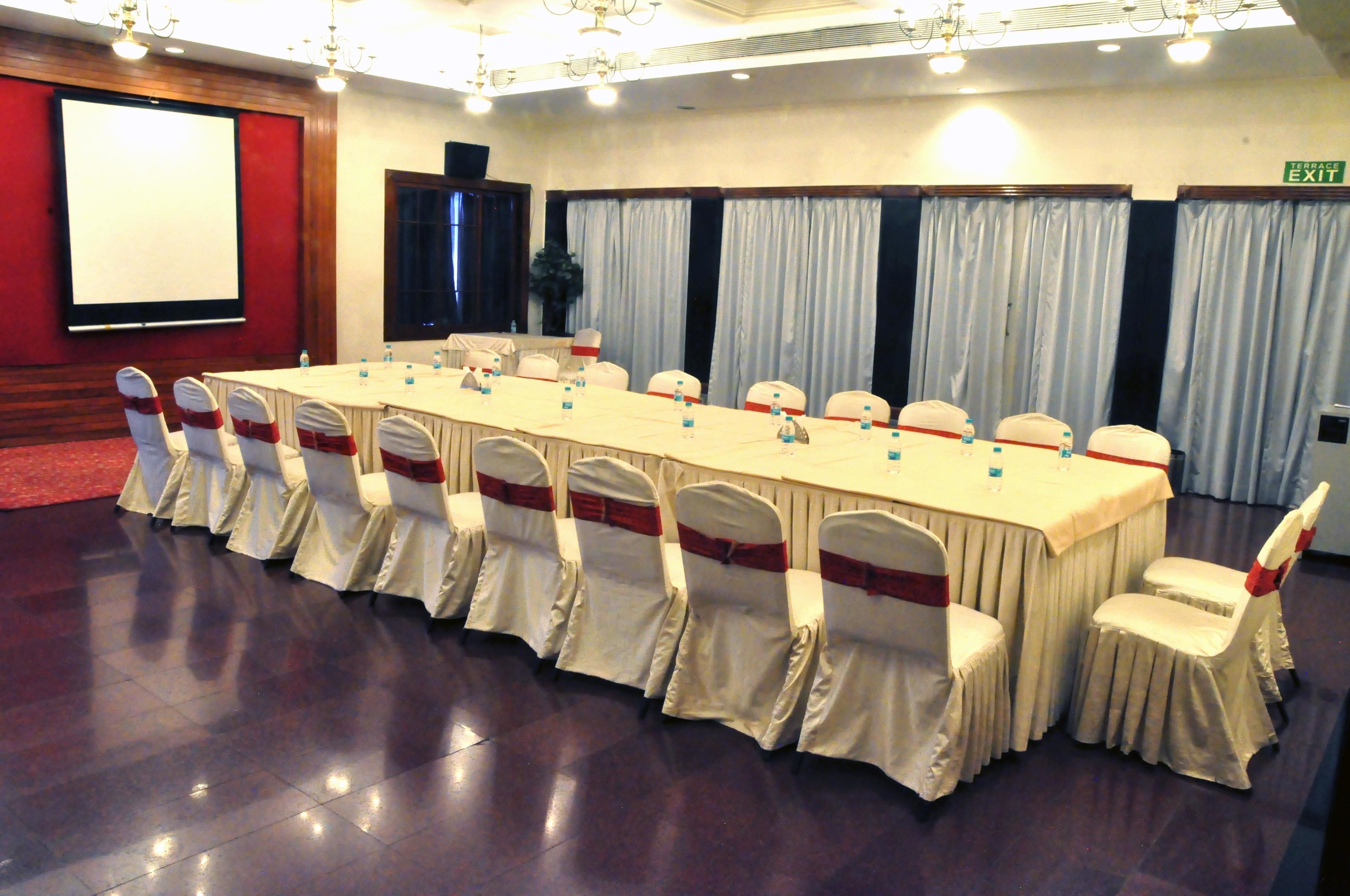 Un salón de conferencias con una larga mesa, sillas con lazos rojos y una pantalla de proyector.