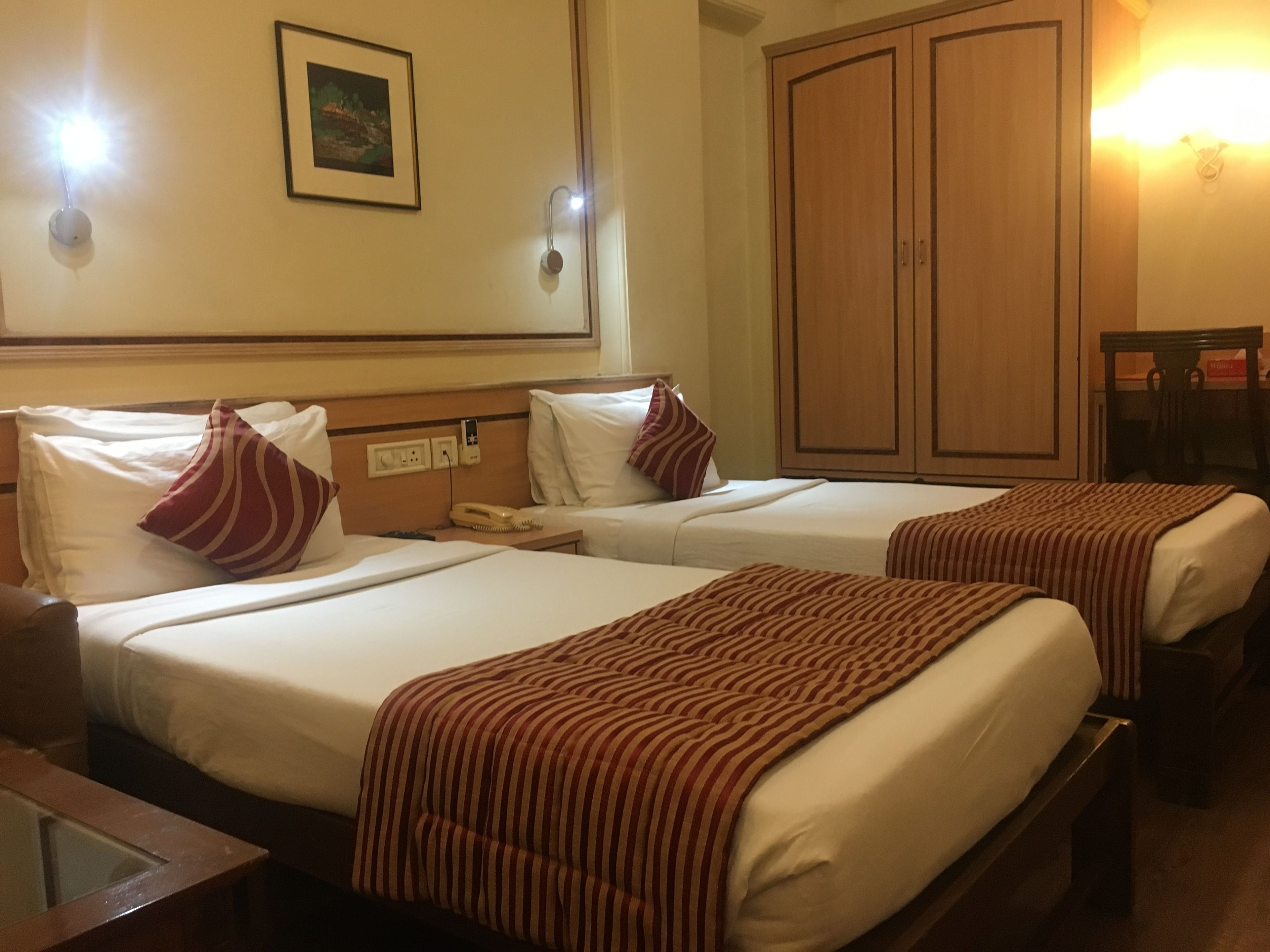 A hotel room with two beds, a wooden wardrobe, a bedside table with a lamp, and a framed picture on the wall.