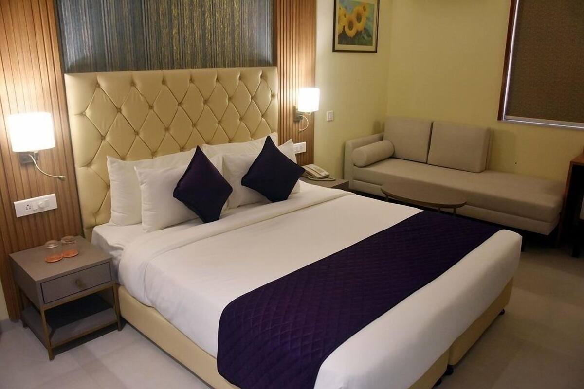 Quarto de hotel com uma cama grande, mesa de cabeceira, sofá e luminária fixada na parede.