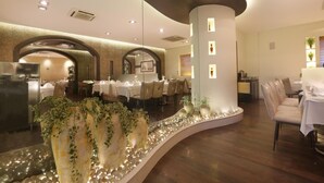 A restaurant with round tables and chairs, a curved wall with decorative plants, and a column with illuminated panels. - Abbott Hotel (Navi Mumbai)