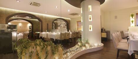 A restaurant with round tables and chairs, a curved wall with decorative plants, and a column with illuminated panels.