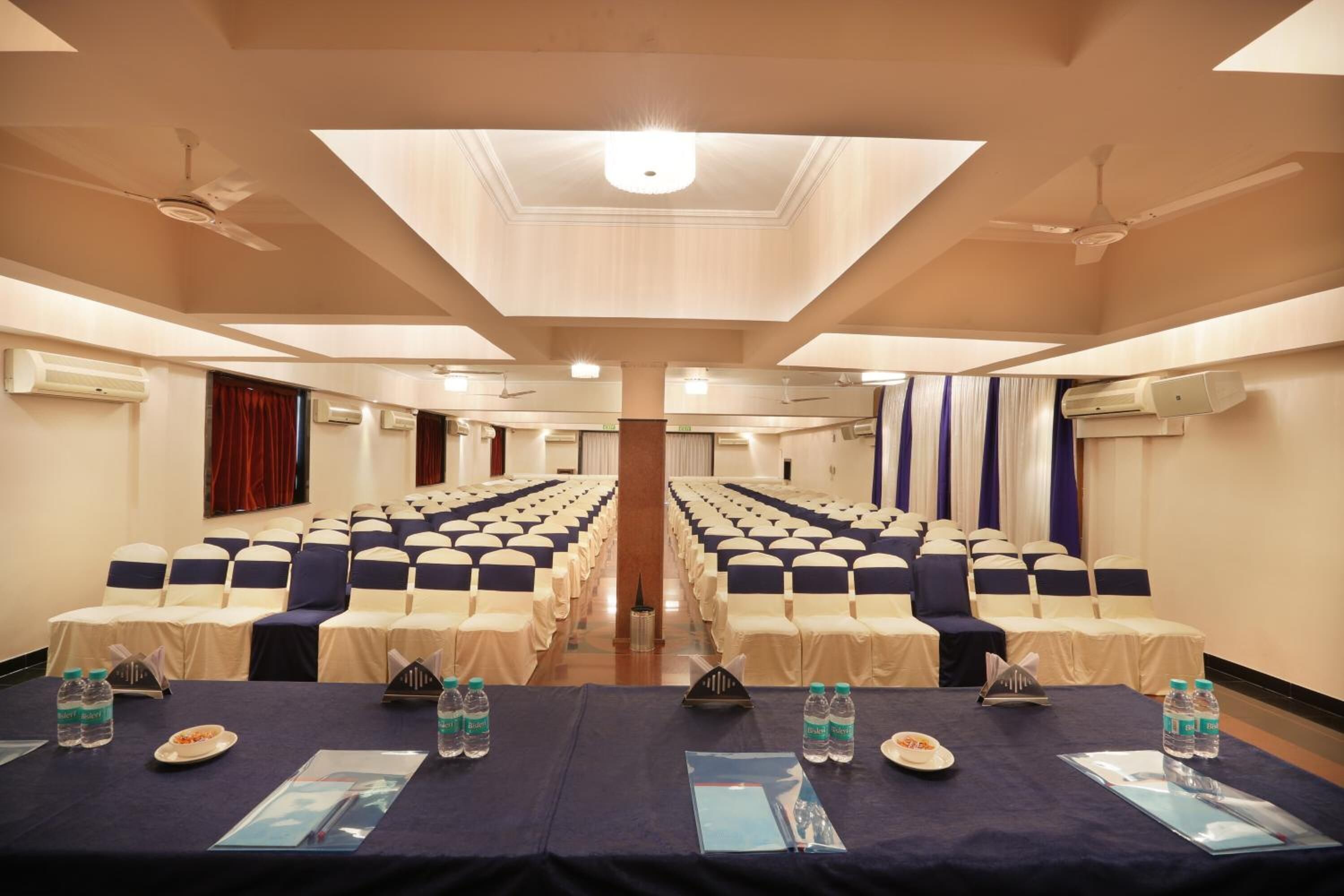 A conference room with rows of chairs, tables with water bottles, and air conditioning units on the walls.