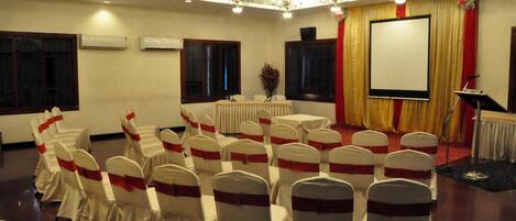 A conference room with rows of white chairs with red sashes, a podium, a screen, and chandeliers.