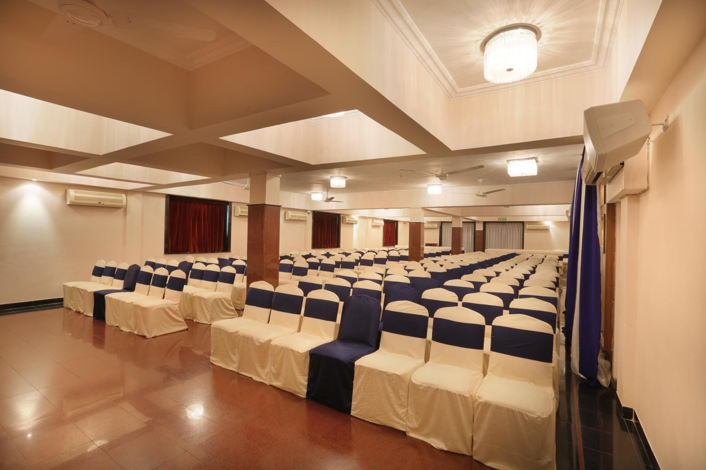 A large hall with rows of chairs and tables, air conditioning units, and a ceiling with recessed lighting.