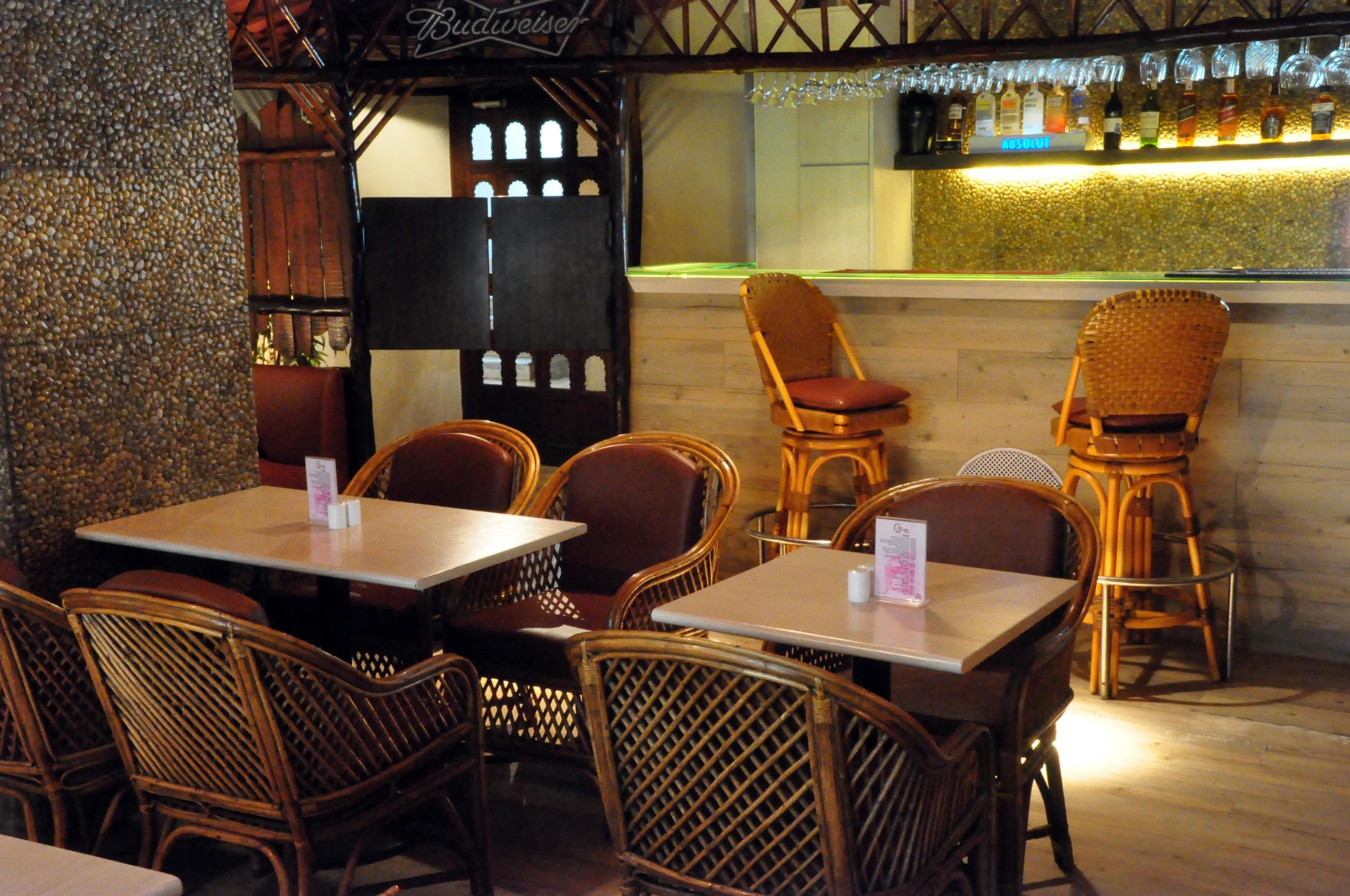 Un restaurante con sillas y mesas de mimbre, una zona de bar con botellas y una pared decorativa.