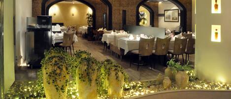 Un restaurante con suelo de madera, mesas y sillas blancas. Hay plantas en macetas grandes y una tira decorativa de luces a lo largo de la pared.