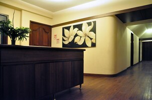 A hallway with a wooden reception desk, a large floral painting on the wall, and a fire alarm.