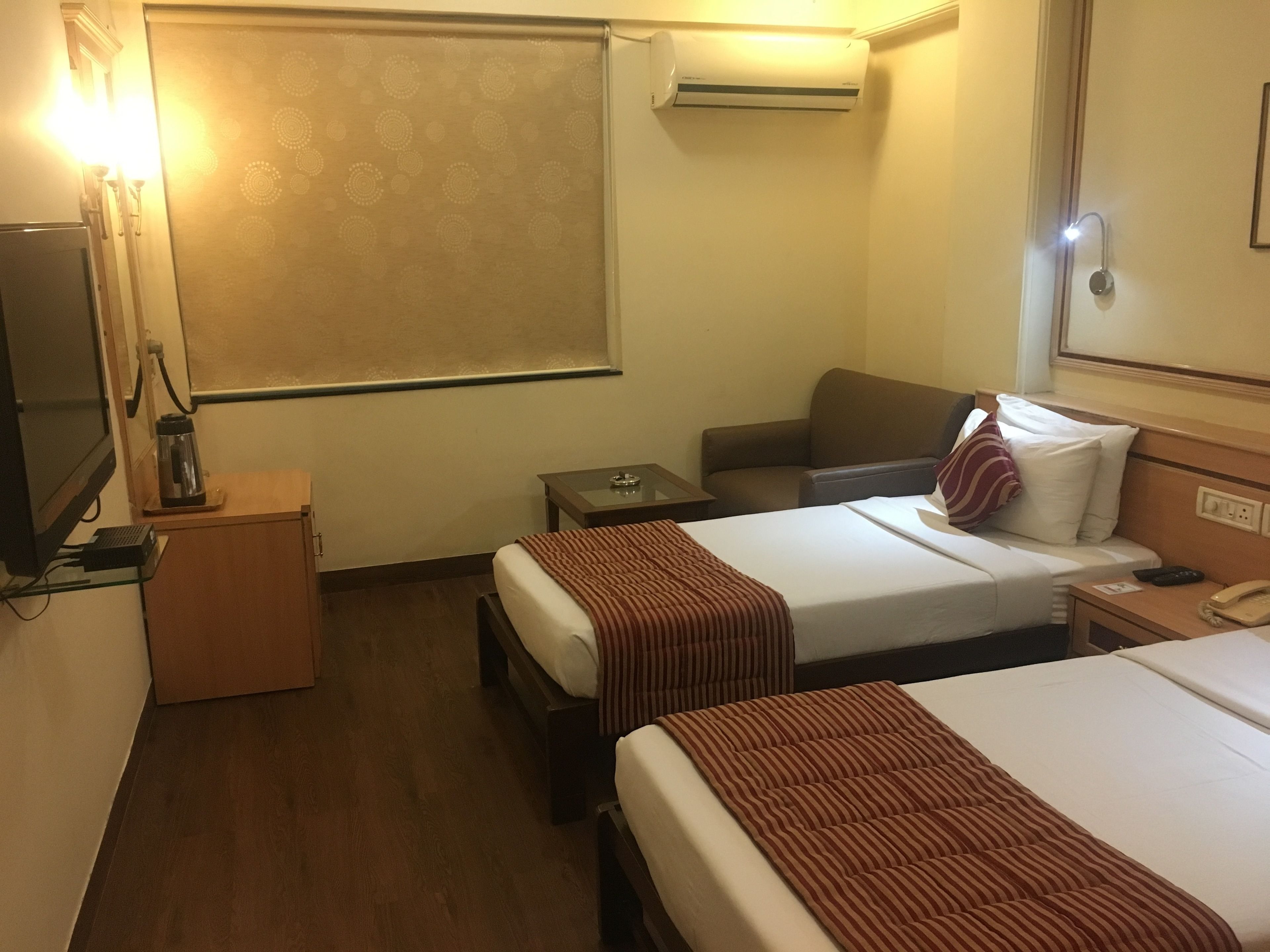 A hotel room with two beds, a bedside table, a nightstand, a lamp, a window with blinds, an air conditioning unit, and a wall-mounted telephone.