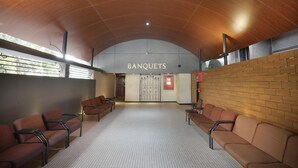 A spacious waiting area with a high wooden ceiling and a patterned floor. There are rows of brown chairs and a sign that reads "BANQUETS". - Abbott Hotel (Navi Mumbai)