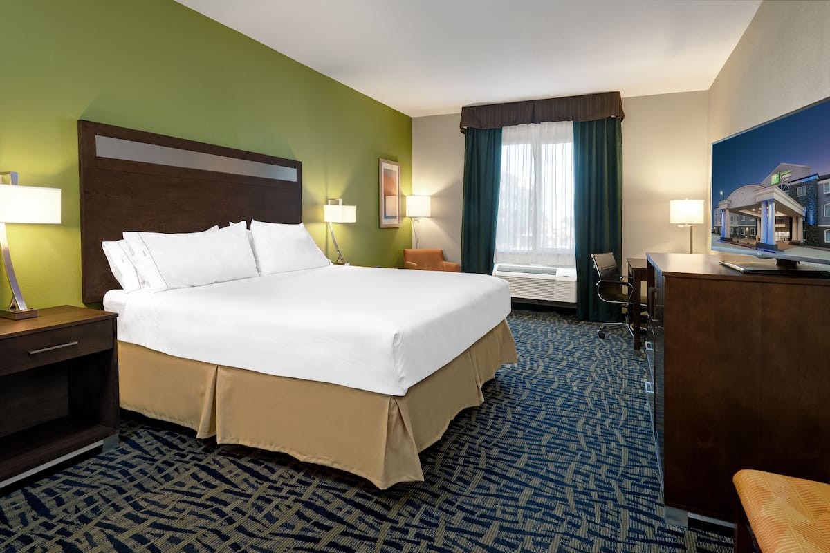 A hotel room with a large bed, two bedside tables with lamps, a desk with a chair, a television, and a window with curtains.