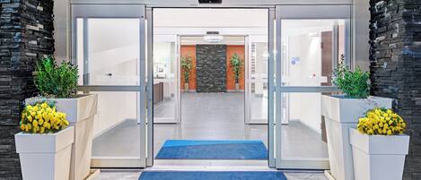 Entrada de hotel con puerta de vidrio, alfombra azul y plantas en macetas a ambos lados.