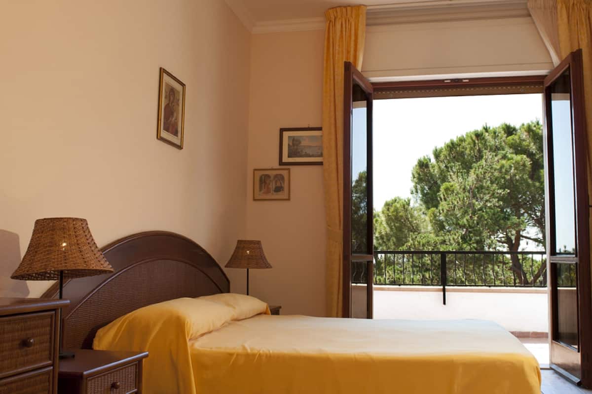A bedroom with a bed, two bedside lamps, a dresser, and a balcony with a view of trees.