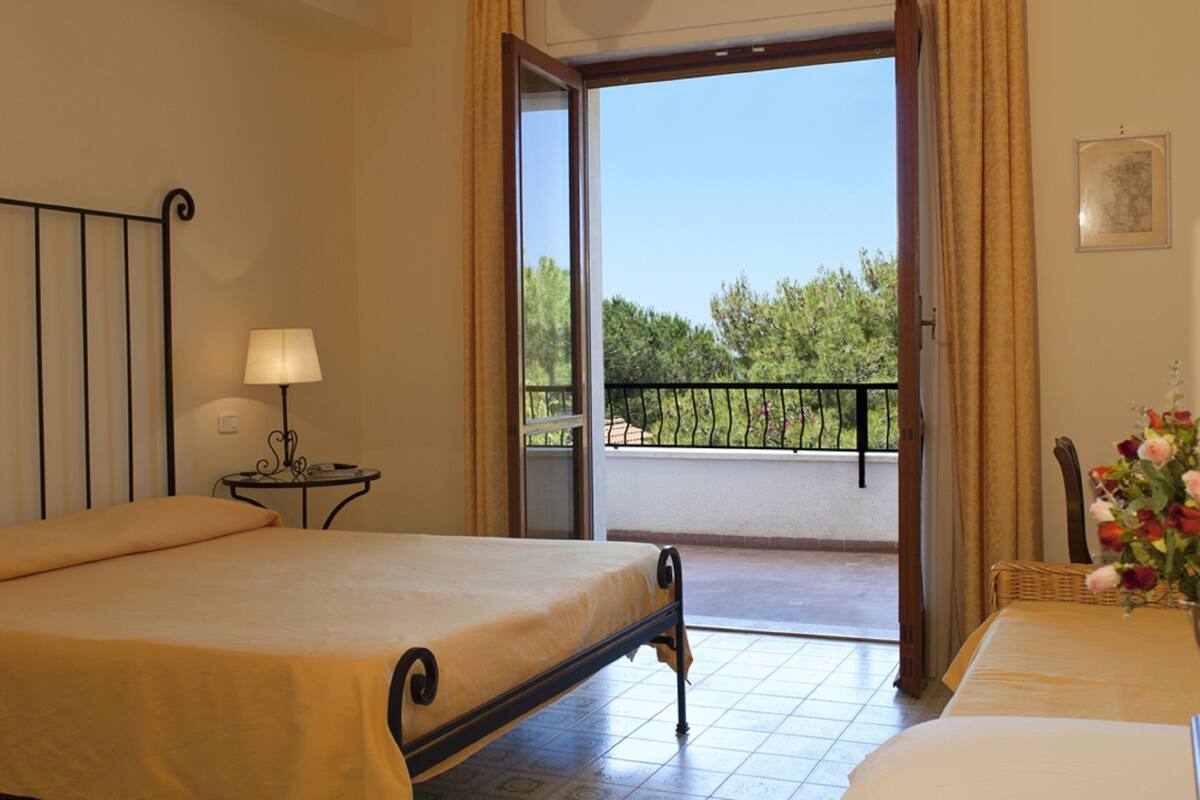 A hotel room with a bed, a small table with a lamp, and a balcony with a view of trees and a fence.